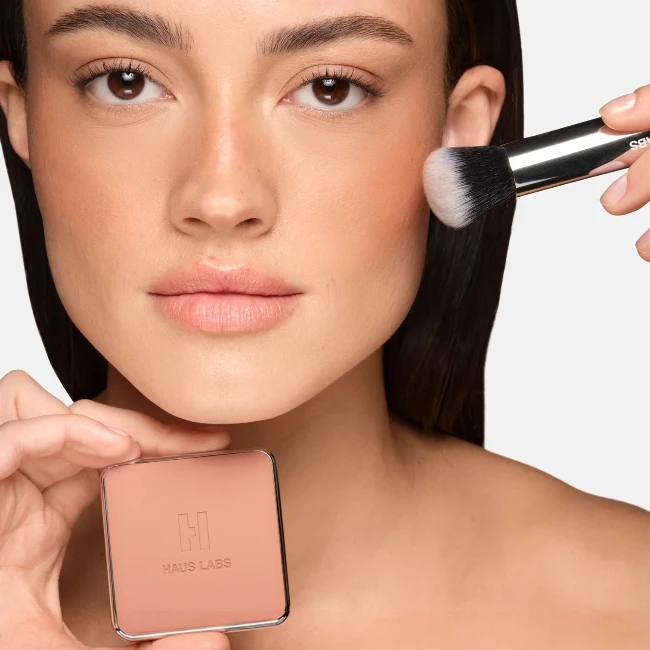 Bir kadın, sağ yanağına sıcak alt tonlu bir allık fırçayla uygularken, sol elinde tuttuğu Haus Labs Color Fuse Blush kompaktı 2026 sonbaharda trend olan allık tonları doğal ve sağlıklı allık tonlarından birini yansıtarak yüze doğal bir kızıllık ve sıcaklık katıyor.
