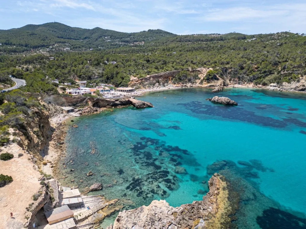 Ibiza'daki yemyeşil tepelerle çevrili, kayalık kıyılara sahip, berrak turkuaz rengi deniziyle dibi görünen, sahilde beyaz binaların ve kıvrımlı bir yolun bulunduğu, birkaç kişinin denizde yüzdüğü ve kumsalda dinlendiği sakin bir koyun kuşbakışı görüntüsü.