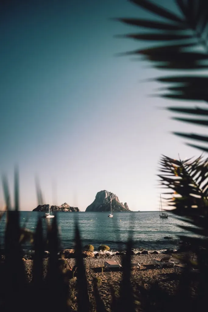 Ibiza'ya ne zaman gidilir sorusuna harika bir cevap sunan bu görüntü, berrak denizde yüzen yelkenliler, uzakta yükselen görkemli kayalık adalar ve ön planda hafifçe görünen plaj bitkileriyle unutulmaz bir tatil anını tasvir ediyor.