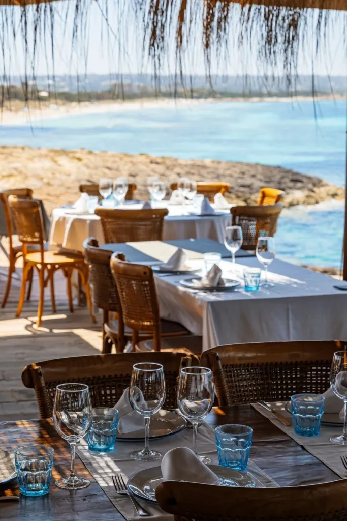 Ibiza'nın masmavi deniz manzarasına sahip, hasır sandalyeler, keten örtülü masalar, zarif kadehler ve canlı turkuaz renkli su bardaklarıyla özenle donatılmış, doğal hasır çatının altında huzurlu bir yemek deneyimi vaat eden bir restoranın iç açıcı detayları.