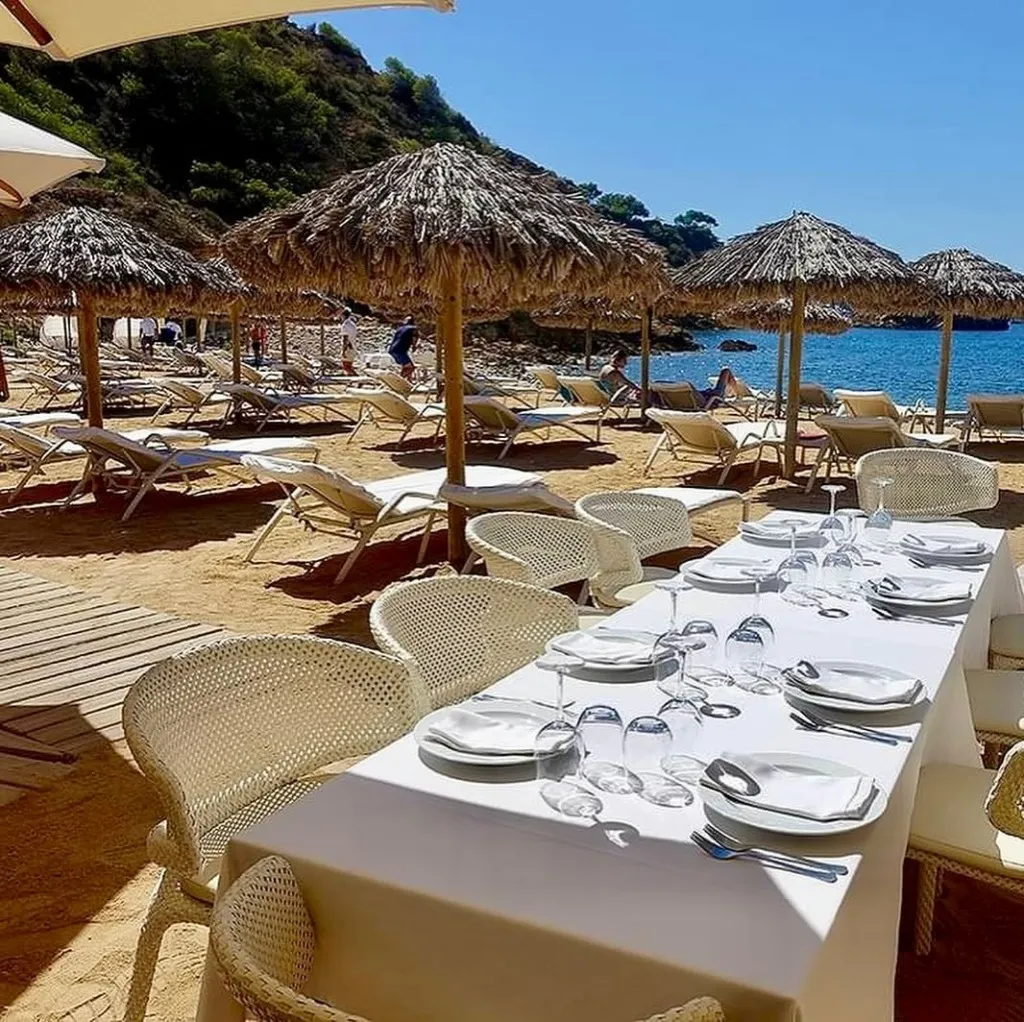 Ibiza'nın kumlu plajında, bembeyaz örtülü, tabak ve kadehlerle özenle hazırlanmış uzun bir yemek masası ile hasır sandalyeler, arka planda güneşlenen insanlar ve masmavi deniz manzarası eşliğinde lüks bir restoran atmosferi.