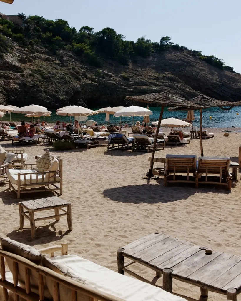 Ibiza'nın güneşli bir plajında, konforlu ahşap şezlonglar ve beyaz plaj şemsiyeleri altında dinlenen insanların bulunduğu hareketli bir manzara; ön planda rustik ahşap masalar ve oturma alanları, arka planda ise berrak turkuaz deniz ve yemyeşil ağaçlarla kaplı kayalık yamaçlar, buranın popüler bir Ibiza restoranı veya plaj kulübü olduğunu düşündürüyor.