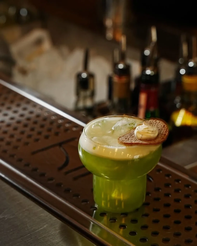 Metal delikli bir bar matı üzerinde, özgün tasarımlı, şeffaf bir bardakta servis edilmiş, buz küpleri ve beyaz köpükle zenginleştirilmiş parlak yeşil bir kokteyl; bardağın kenarına özenle yerleştirilmiş, üzerinde kremalı bir dip sosu olan küçük, koyu renkli bir ekmek dilimi ile tamamlanıyor; arka planda bulanık bar şişeleri seçilebiliyor.