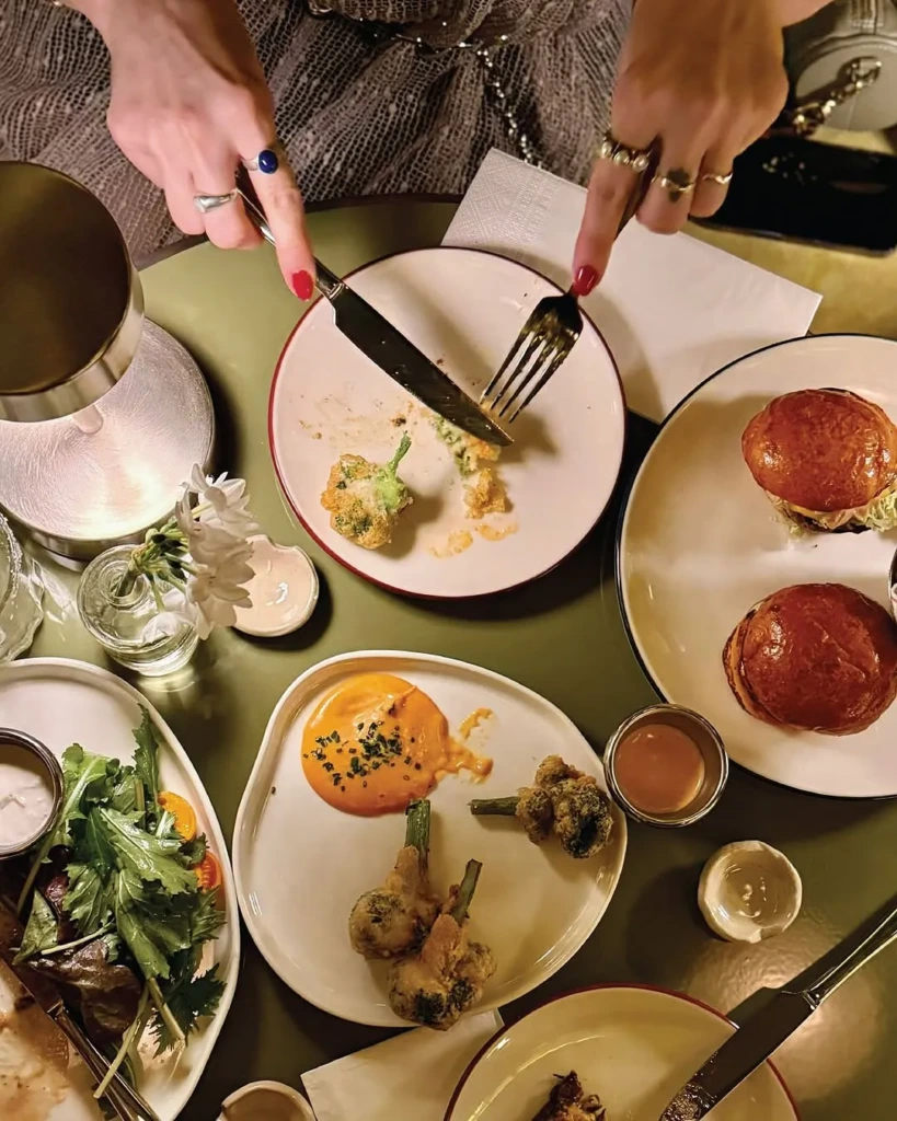 Kiff Bebek restoranında çekilen bu üstten görünüm fotoğrafında, kırmızı ojeli ve yüzüklü ellerin yeşil bir masada, kızarmış brokoli tempurayı bıçak ve çatalla kestiği an net bir şekilde görülüyor; masada ayrıca salata, soslu brokoli ve bürgerler gibi çeşitli özel lezzetler iştah açıcı bir şekilde sergileniyor.