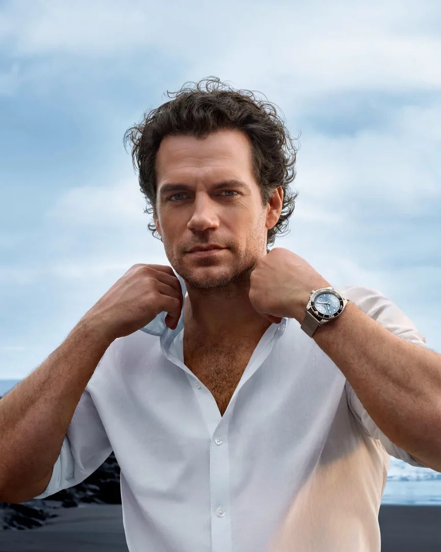 Ünlü oyuncu Henry Cavill, beyaz açık yakalı gömleğinin yakalarını tutarak, sol bileğinde mesh kayışlı, mavi kadranlı zarif bir Longines saat taşıyor, dağınık saçları ve hafif sakallarıyla sahilde objektife bakıyor.