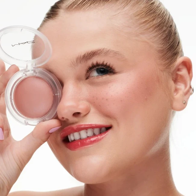 2026 sonbaharında trend olan allık tonlarını yansıtan, gülümseyen genç bir kadının yanaklarındaki doğal çilleri ve pembe parlak dudaklarıyla birlikte, MAC Glow Play Blush kompaktını gözünün yanında tutarak ışıltılı ve doğal bir makyaj görünümü sergilediği yakın çekim editoryal güzellik fotoğrafı.