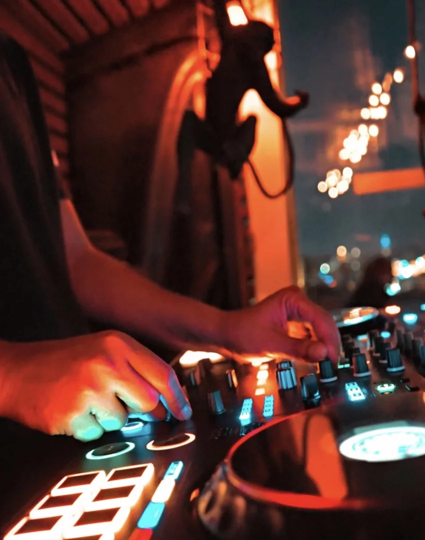Gece atmosferinde, elleri parlak mavi, turuncu ve beyaz ışıklarla aydınlatılmış bir DJ kontrolcüsünü kullanan bir kişinin yakın çekim görüntüsü, arka planda kırmızı renkte aydınlatılmış maymun figürü ve bulanık şehir ışıklarının eşlik ettiği dinamik bir gece kulübü veya bar ortamını gözler önüne seriyor.