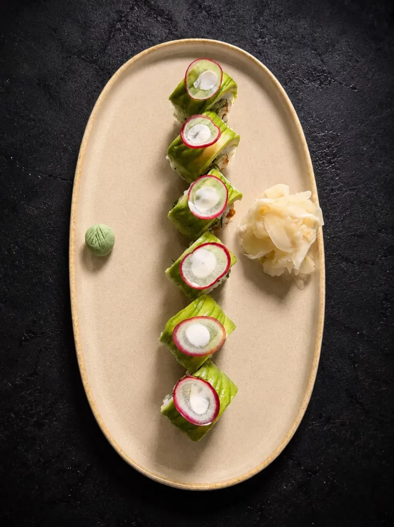 Bej oval bir tabağın üzerinde koyu renkli bir zeminde, dikey olarak sıralanmış altı adet avokado kaplı sushi parçası görünüyor. Her sushi rulosunun üzerinde ince bir turp dilimi ve ortasında beyaz bir krema bulunurken, tabağın yanında küçük bir top yeşil wasabi ve bir yığın dilimlenmiş zencefil turşusu yer alıyor.