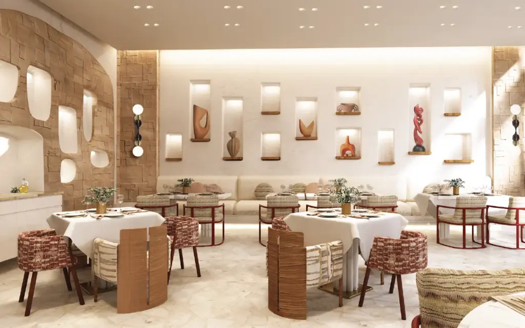 Aydınlık ve modern bir restoranın içinde, taş dokulu kemerli bir duvar ile nişlerde sergilenen toprak tonlarında soyut heykellerin ve bir kırmızı heykelin bulunduğu beyaz bir duvarın ortasında, beyaz masa örtülü yemek masaları ve desenli, özgün sandalyelerle zarif bir oturma düzeni görülüyor.