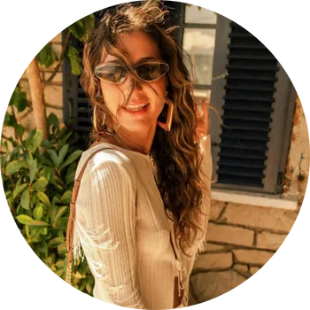 Güneş gözlüğü ve büyük küpeler takmış, uzun dalgalı saçları rüzgarda hafifçe dağılmış, yırtık detaylı şık beyaz ribana bluzuyla kamera gülümseyen Narod Çetinkaya, arkasında taş duvar ve yeşil bitkilerin olduğu sıcak bir dış mekanda poz veriyor.