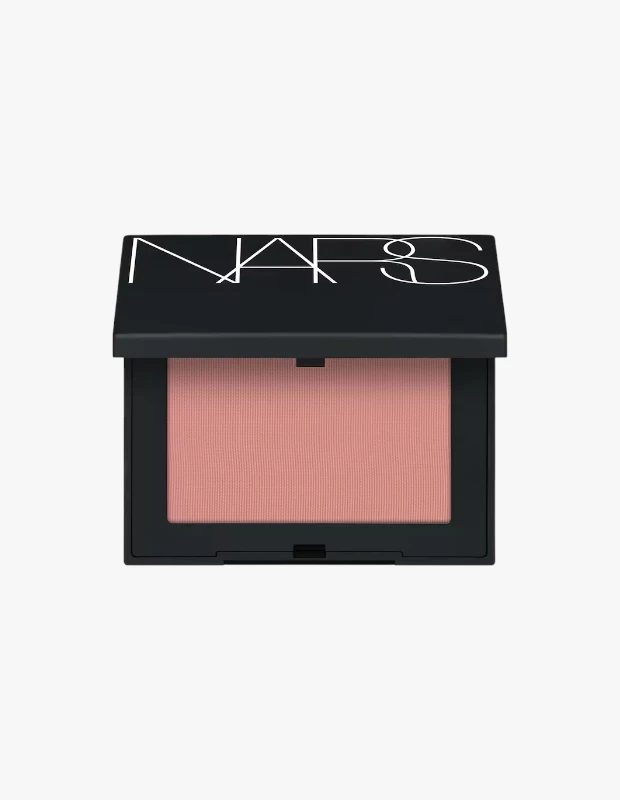 NARS markalı, kapağı hafifçe açık duran mat siyah kompakt kutusunda görünen, yumuşak dokulu açık şeftali pembe Behave rengi talksız pudra allık, hassas ciltler için uygun kozmetik ürünlerinin önemini vurguluyor.