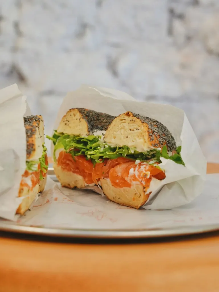 Gümüş bir tepside, beyaz ambalaj kağıdına sarılı, üzerine haşhaş tohumlarıyla süslenmiş, ortadan ikiye kesilmiş bir bagel sandviçinin içinden bol miktarda narince dilimlenmiş füme somon, krem peynir ve canlı yeşil roka yaprakları taşıyor; bu keyifli ve iştah açıcı lezzet, arka plandaki bulanık beyaz tuğla duvar önünde sunuluyor.