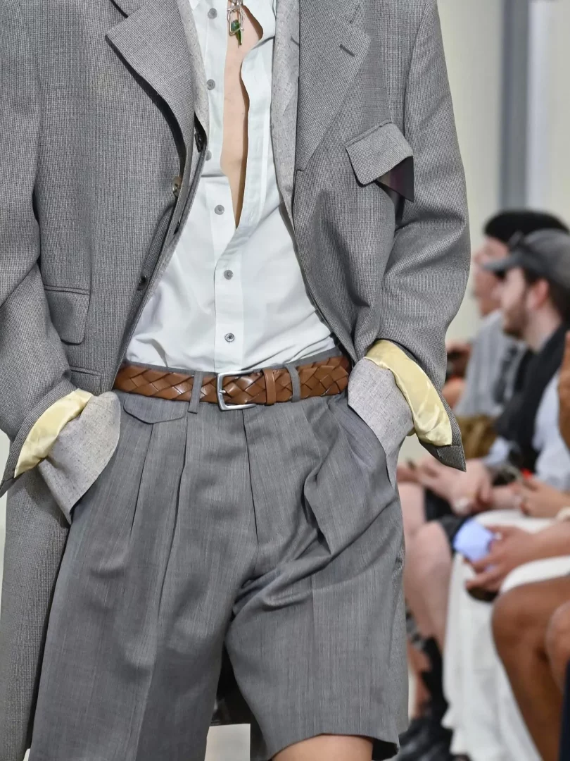 Paul Smith defilesinden detay bir karede, açık renk gömlek, gri blazer ceket ve şort kombini giyen bir erkek model, kahverengi örgülü deri kemer ve boynundaki yeşil taşlı kolyesiyle şık bir Paul Smith erkek aksesuar görünümü sunuyor.