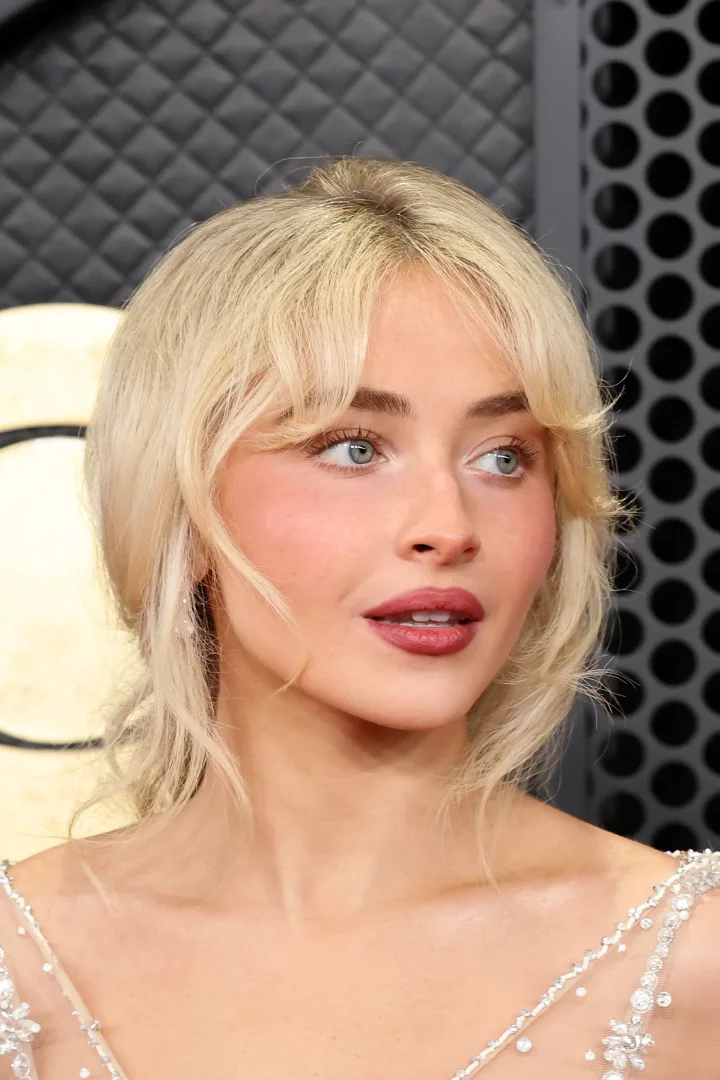 Sabrina Carpenter, sarı dalgalı saçları, belirgin kirpikleri ve pembe yanaklarıyla dikkat çeken bebeksi göz makyajını sergilerken, parıltılı işlemeli bir elbiseyle koyu renk, dokulu bir arka plan önünde poz veriyor.