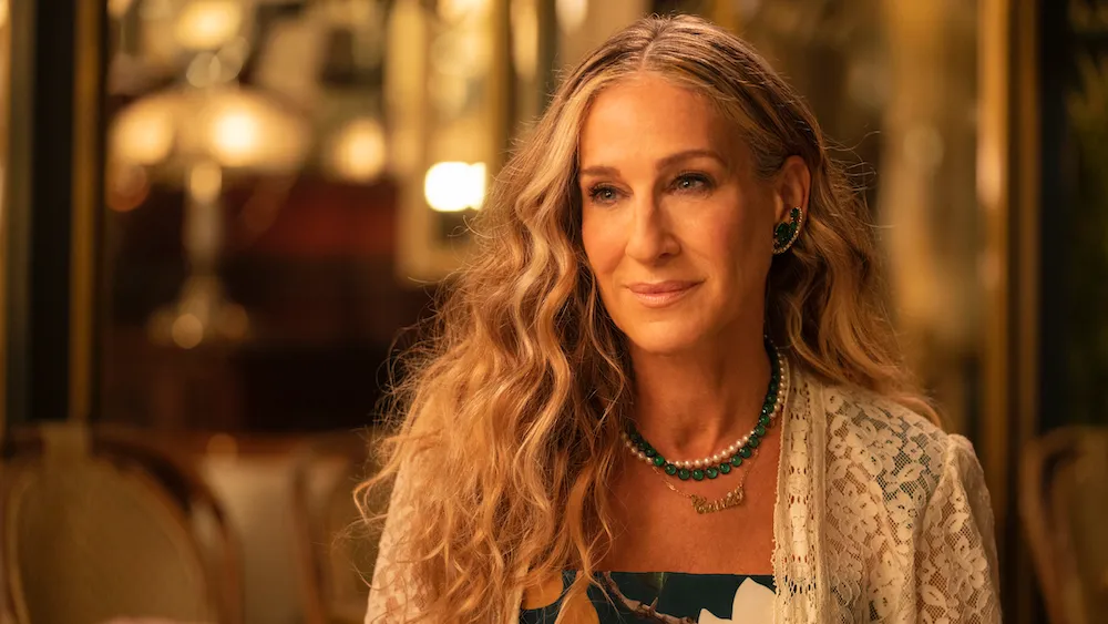 Sarah Jessica Parker'ın uzun dalgalı sarı-gri saçları, yeşil taşlı küpeleri ve inci ile yeşil boncuklu iki katmanlı kolyeleriyle hafif bir gülümsemeyle kameraya hafifçe yandan bakarak zarif bir şekilde poz verdiği yakın çekim portresi, arkasında bulanık sıcak ışıklar ve mobilya detayları seçilen lüks bir iç mekanda çekilmiş.