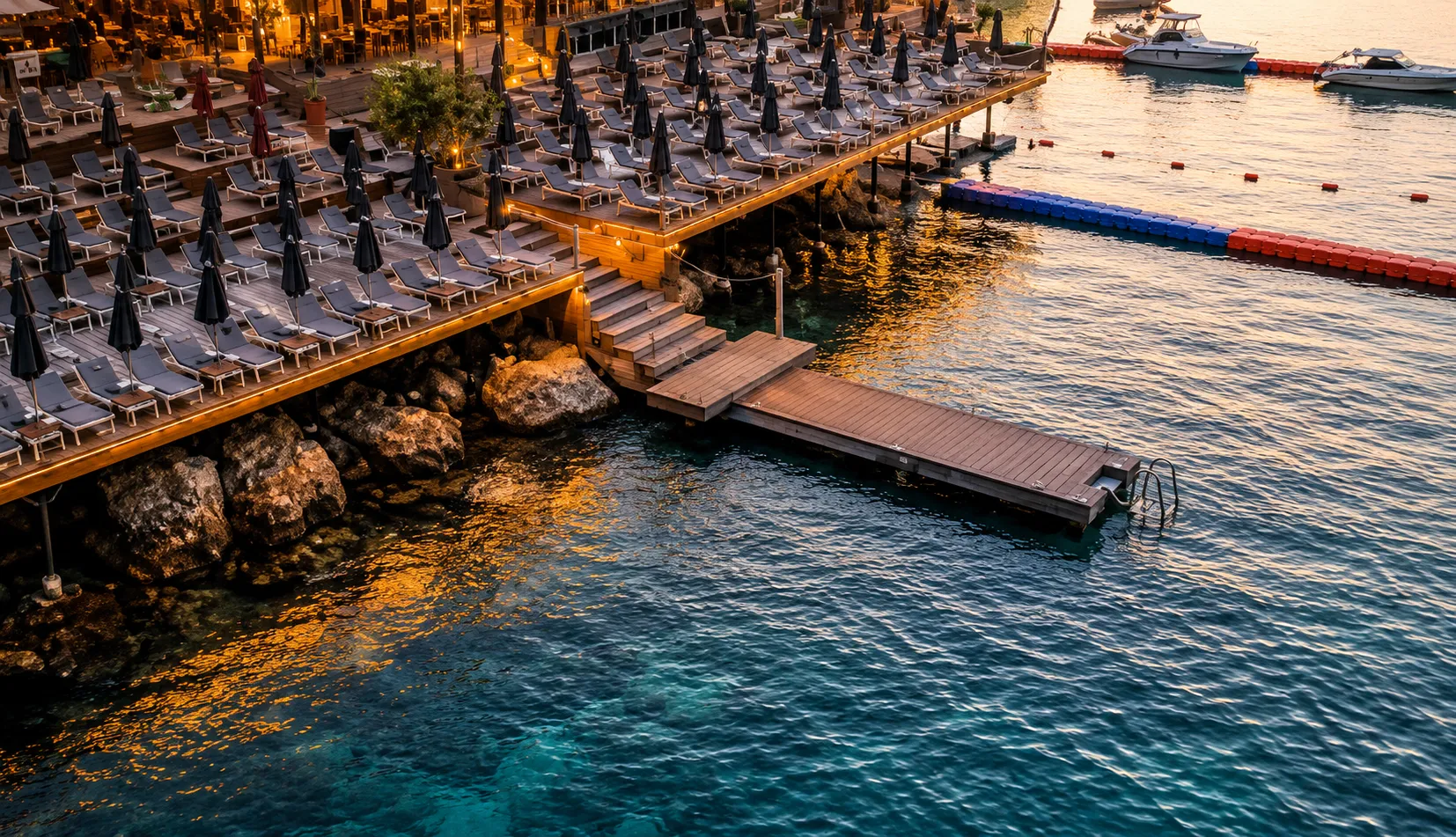 Sarnıç Beach Club Bitez’de ahşap teras, şezlonglar, iskele ve gün batımında sakin deniz manzarası