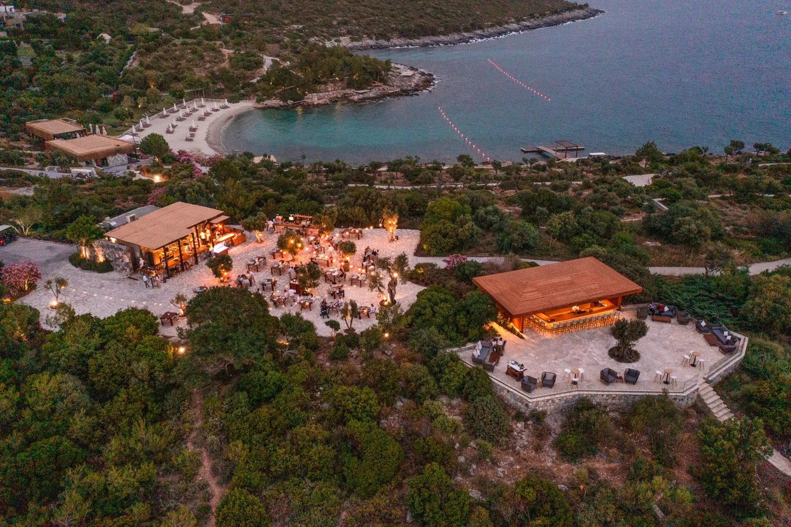 Ege kıyısında gün batımında çekilmiş havadan görünüm; yemyeşil doğanın içinde konumlanan, taş teraslı ve ahşap çatılı yapıların bulunduğu lüks resort alanı, aşağıda sakin koy ve turkuaz deniz manzarasıyla birlikte.