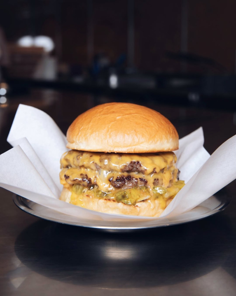 Galataport'un öne çıkan mekanlarından Akali'de servis edilen, parlak hamburger ekmeği arasında bol erimiş cheddar peyniri ve turşularla zenginleştirilmiş, iki katlı sulu et köftesiyle son derece iştah açıcı ve doyurucu bir çift burgerin yakından çekilmiş fotoğrafı.