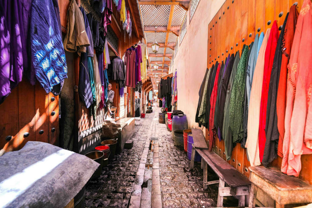 Marakeş'in daracık, Arnavut kaldırımlı bir çarşısında, ahşap dükkan cepheleri boyunca asılı mor, mavi, kırmızı ve yeşil tonlarda binbir çeşit rengarenk geleneksel kıyafetler ve yansıyan ışık oyunlarıyla dolu canlı bir alışveriş atmosferi tasvir ediliyor.