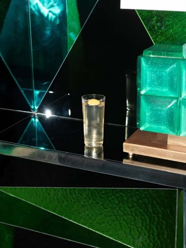 Limon dilimli bir içeceğin, yansıtıcı koyu bir yüzeyde konumlandığı, arka planda zümrüt yeşili cam küpler ve geometrik panellerle çevrili, modern ve sofistike bir bar veya mekan detayı.