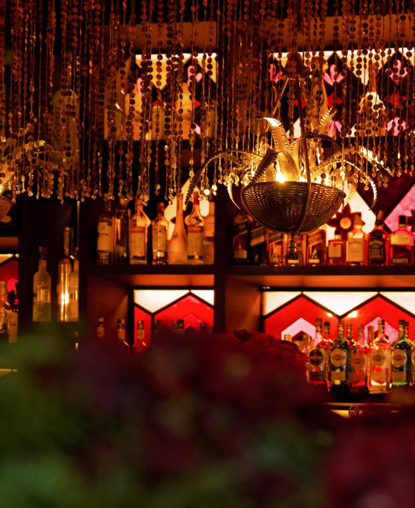 Marakeş’in en iyi restoranları arasında yer alan Comptoir Darna’nın, arka planda sıralanmış içki şişeleri ve kırmızı tonlu panellerle vurgulanan egzotik bar alanını gösteren, tavanından sarkan parlak altın boncuklu perdeler ve ortasında metalik süslemeli bir avizenin loş ışıklar altında büyüleyici bir atmosfer yarattığı detaylı bir görüntü.