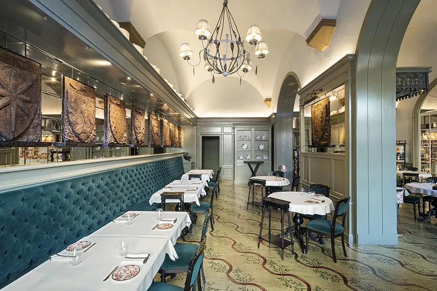 Mevcut görselde, Floransa'daki lüks Gucci Osteria restoranının şık iç mekanı görülüyor; uzun, kadife yeşili banket oturma düzeni, beyaz örtülü masalar, zarif desenli yer döşemeleri ve duvarları süsleyen kabartma paneller, mekanın sofistike atmosferini vurguluyor.