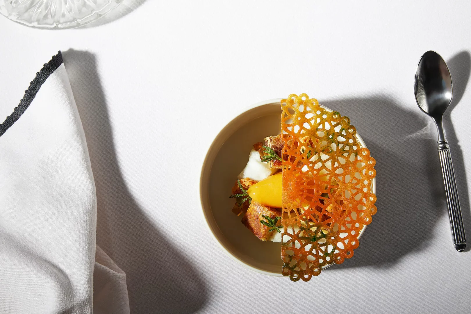 Beyaz masa örtüsü üzerinde, Gucci Osteria'nın yaratıcı mutfağından eşsiz bir sunum: turuncu dantel görünümlü bir çıtırla süslenmiş, parlak sarı bir püre, lezzetli et parçaları ve taze yeşil otlarla bezenmiş, zarif bir kasede servis edilen gurme bir yemek, yanında gümüş bir kaşık ve kenarı siyah işlemeli beyaz bir peçete ile detaylandırılmış genel görünüm.