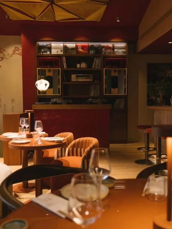 Koyu kırmızı duvarların önünde, plaklar ve ses sistemleriyle dolu özel bir nişin de bulunduğu, sıcak ışıklandırılmış modern restoranda, şık bir şekilde düzenlenmiş yemek masaları ve kadife görünümlü turuncu sandalyeler dikkat çekiyor.