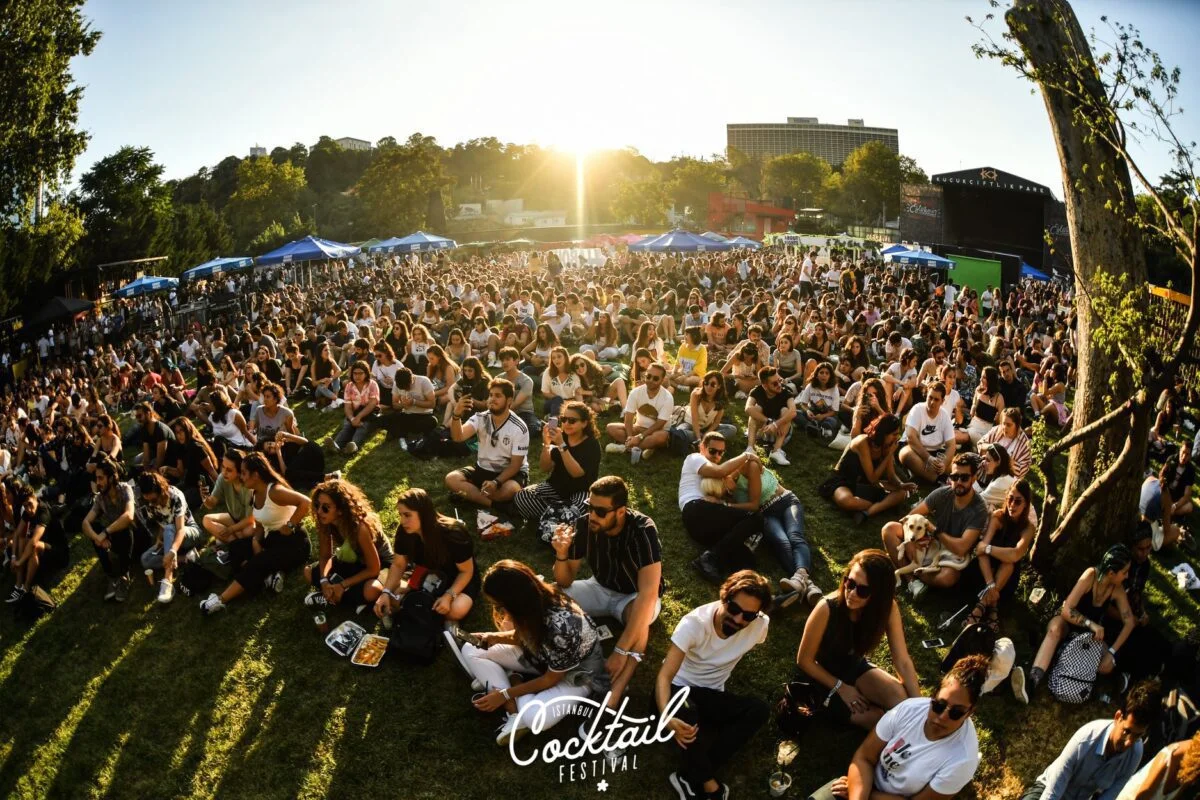 İstanbul Cocktail Festival'inde gün batımının sıcak ışıkları altında çimlerde oturan büyük bir kalabalık, müziğin ve kokteyl kültürünü keşfetmenin keyfini çıkarırken şehrin sosyal hayatına canlı bir enerji katıyor.