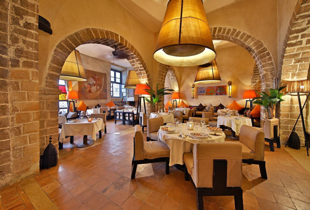 Marakeş’in en iyi restoranları arasında dikkat çeken La Table by Madada’nın, tuğla kemerlerle zenginleşen taş duvarları, sıcak sarı ışık veren devasa kumaş avizeleri ve portakal renkli lambalarıyla aydınlatılmış, zarif beyaz masa örtülü yuvarlak masalar ve konforlu koltuklarla dolu samimi yemek salonu.