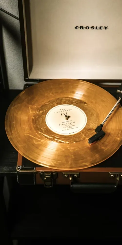 CROSLEY marka taşınabilir bir pikapta çalan, üzerinde The Lumineers III yazılı etiketiyle dikkat çeken, parıltılı altın rengi bir plağın yakın çekim görüntüsü, müzikseverler arasında neden bu kadar popüler olduğunu gösteren bir listening room deneyimini gözler önüne seriyor.