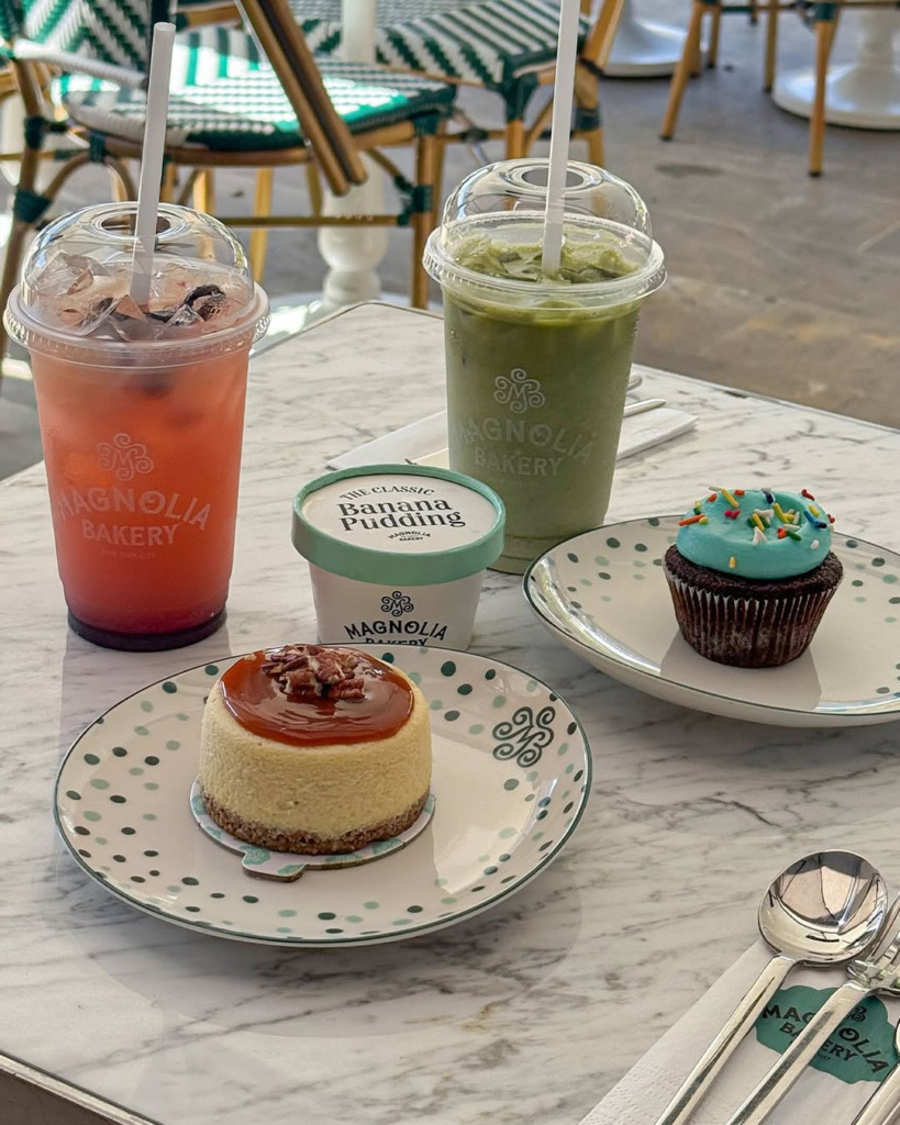 Galataport'un tatlı severler için en iyi mekanlarından Magnolia Bakery'de, karamelli pekanlı cheesecake, mavi kremalı renkli şekerlemeli cupcake, muzlu puding ve iki farklı buzlu içeceğin mermer bir masada şık sunumu görülüyor.