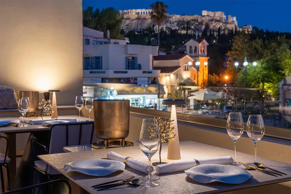 Atina'daki lüks bir restoranın terasında, masalar şık bir şekilde hazırlanmış; beyaz tabaklar, kadehler ve modern masa lambalarıyla donatılmışken, arka planda Akropolis'in gece ışıklarında büyüleyici silueti ve şehrin ışıltılı binaları görülüyor.