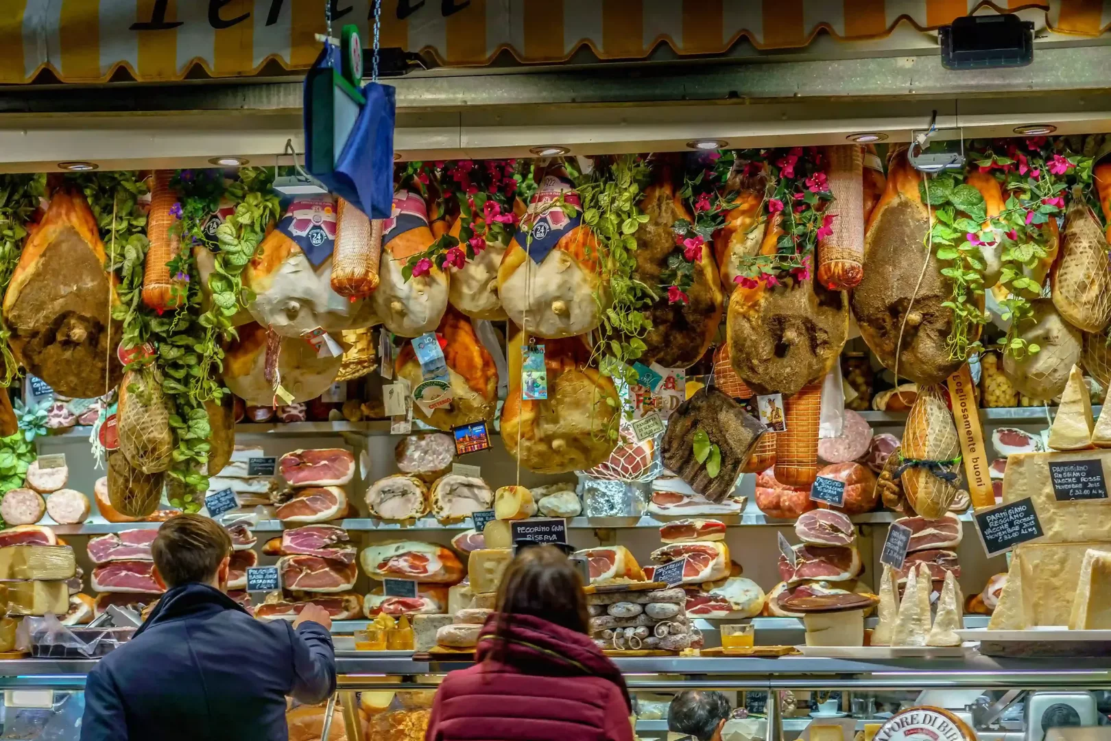 Mercato Centrale'deki bir şarküteri tezgahında, yeşil sarmaşıklar ve pembe çiçeklerle süslenmiş tavanlardan sarkan çok sayıda jambon ve salam çeşidi ile alt raflarda dilimlenmiş etler ve çeşitli peynirler sergilenirken, iki müşteri ürünleri inceliyor.