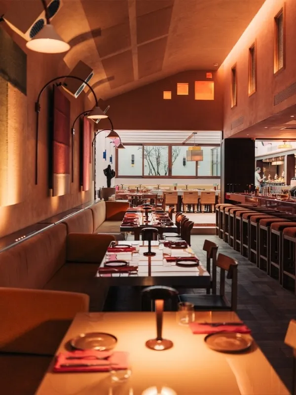 Sıcak ve şık bir restoranın iç mekanı; uzun banket oturma alanında modern lambalarla donatılmış masalar, uzakta bir bar bölümü ve dışarıyı gösteren büyük pencereler ile davetkar bir ambiyansta aydınlatılmış.
