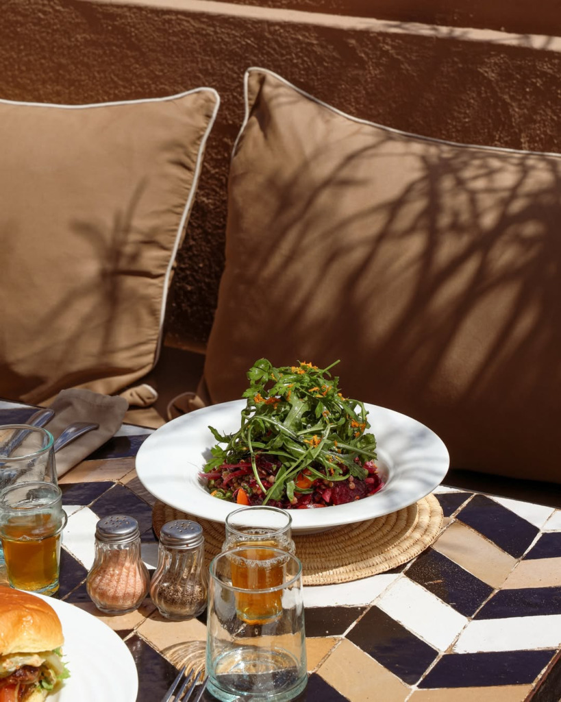 Marakeş'in en iyi restoranlarından Nomad'ın terasında, geometrik desenli fayans masada sunulan bol yeşillikli taze salata ve içecekler, güneşin altında kahverengi minderlerin gölgelerle süslendiği huzurlu bir atmosferde keyifli bir yemek deneyimi vaat ediyor.
