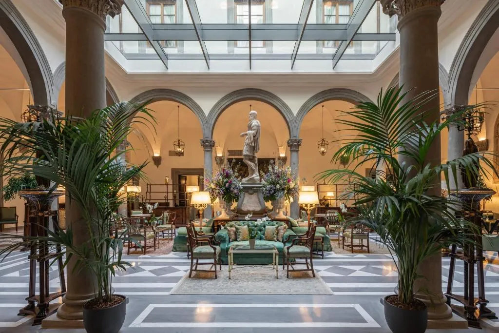 Palazzo Portinari Salviati'nin cam tavanlı geniş avlusunda, klasik bir heykelin ve canlı çiçek düzenlemelerinin ortasında, sütunlar ve kemerlerle çevrili mermer zeminde yeşil koltuk takımları ve palmiye bitkileriyle zenginleştirilmiş lüks bir oturma alanı.
