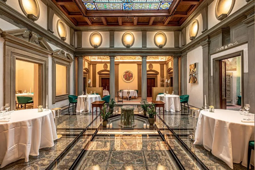 Palazzo Portinari Salviati'nin bu görkemli iç mekanında, cam bir zeminin altında görünen mozaik desenli döşemeler, ortada bitkilerle çevrili bir fıskiye, beyaz örtülü masalar ve yeşil sandalyelerle zenginleştirilmiş lüks bir yemek salonu atmosferi vitray tavanla aydınlanıyor.