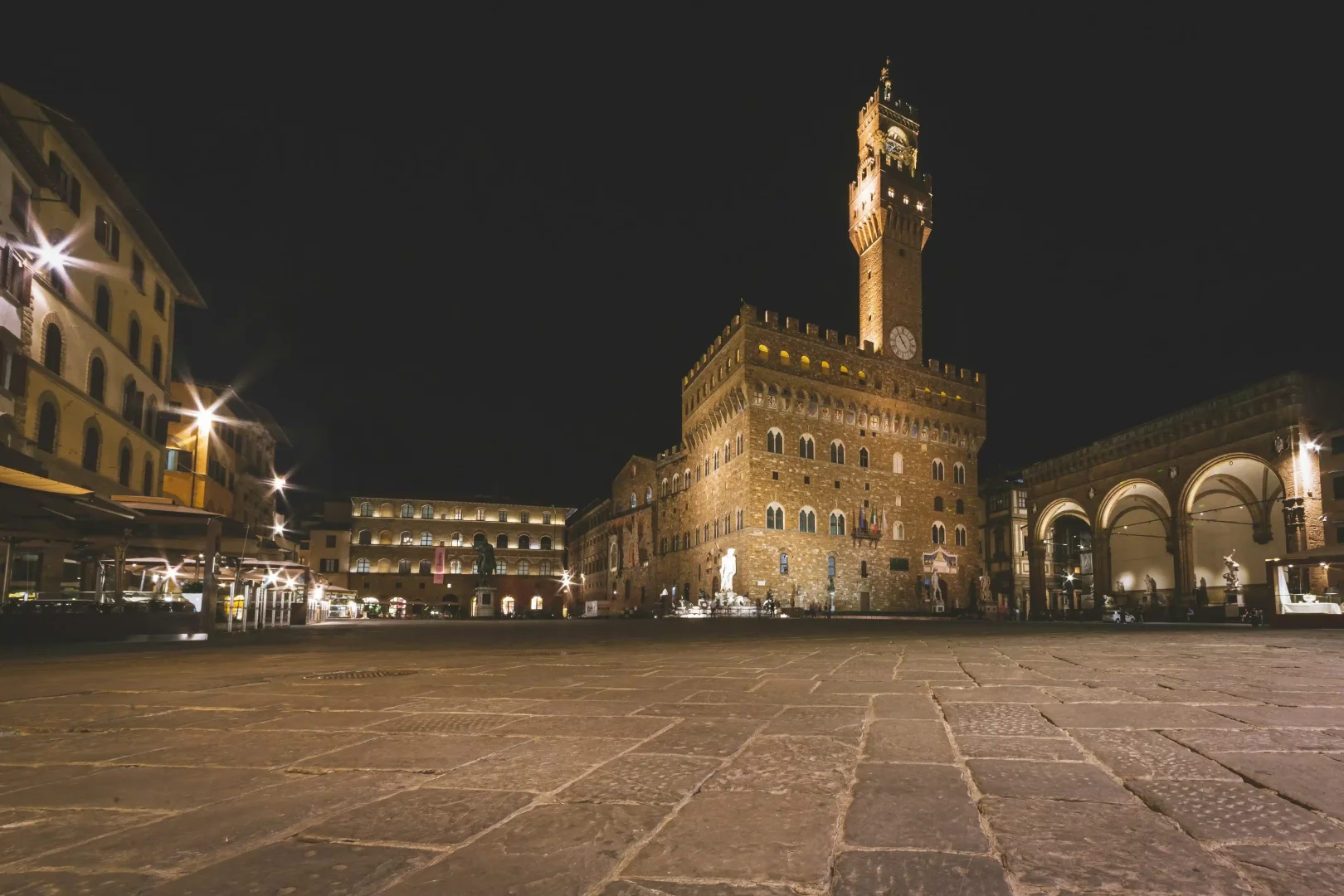 Karanlık gökyüzünün altında, tüm ihtişamıyla ışıklandırılmış, görkemli saat kulesiyle öne çıkan tarihi Palazzo Vecchio'nun, Firenze'nin Signoria Meydanı'ndaki geniş taşlı avlusundan çekilmiş atmosferik bir gece görüntüsü, çevresindeki loş ışıklı binalar ve sağdaki kemerli Loggia dei Lanzi'deki heykellerle beraber sakin ve etkileyici bir an sunuyor.