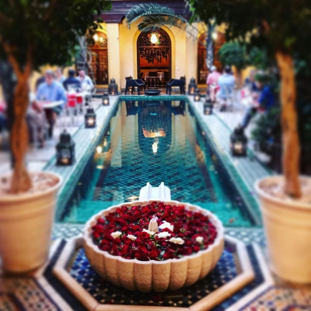 Marakeş Medina'sının kalbindeki Pepe Nero restoranının avlusunda, ortasında kırmızı güller ve küçük bir fıskiye bulunan taş bir kase ile mozaik desenli bir zemine sahip, yansımalı havuzun çevresinde, loş ışıklar ve zarif oturma alanlarıyla lüks bir akşam yemeği atmosferi sunuluyor.