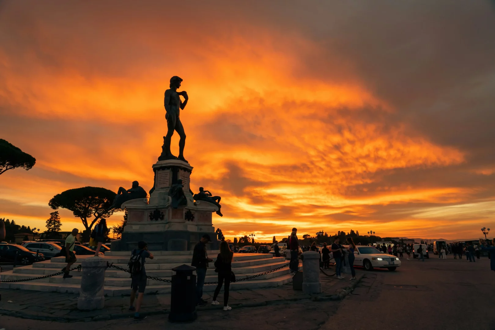 Piazzale Michelangelo'daki Michelangelo'nun Davut heykelinin bronz replikası, etrafında dolaşan insanların ve ağaçların siluetlerini çizen, turuncu ve sarı tonlardaki dramatik gün batımı gökyüzünün altında etkileyici bir görünüm sergiliyor.