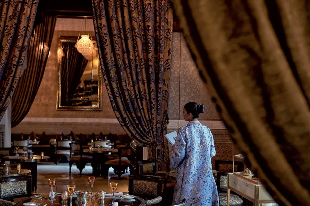 Marakeş'in en iyi restoranlarından Royal Mansour'daki La Grande Table Marocaine'ın zengin ve zarif atmosferinde, işlemeli açık mavi kaftanıyla bir garson, desenli perdelerin ve amber kadehlerle donatılmış lüks masaların arasındaki loş ışıklı salonda hizmete hazır bekliyor.