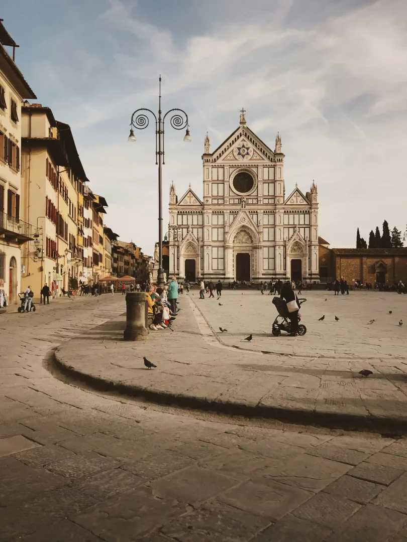 Santa Croce Bazilikası'nın karmaşık desenli beyaz ve yeşil mermer cephesi, önündeki tarihi Floransa meydanında yürüyen ve oturan insanlarla, etrafta uçuşan güvercinlerle birlikte sakin bir günü yansıtan geniş bir görüntüdür.