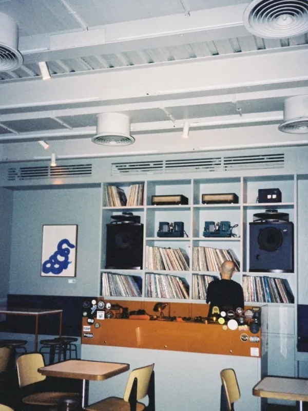 Beyoğlu'ndaki bir taproom'un iç mekanında, geniş bir plak koleksiyonu ve DJ ekipmanlarıyla dolu rafların önünde sırtı dönük bir DJ'in müzik yaptığı, turuncu bir bar tezgahı ve şık oturma alanlarının bulunduğu canlı bir atmosfer yansıyor.