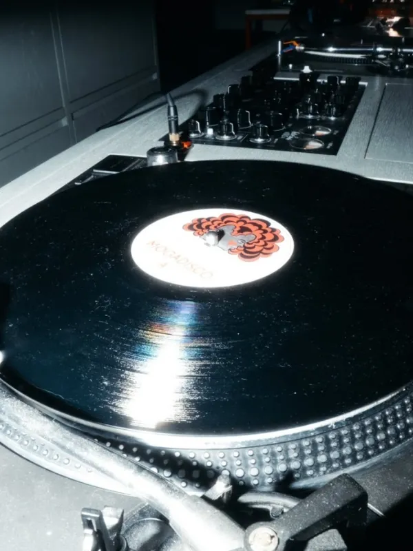 Gece kulübü veya canlı müzik mekanı atmosferini yansıtan loş bir ortamda, kırmızı ve siyah bir desenin olduğu beyaz etiketli parlak siyah bir plak, detaylarıyla görülen bir döner tablada çalmaya hazır duruyor.