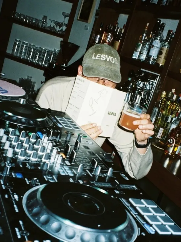 Ön planda bir DJ mikseri bulunurken, yeşil LESVOS şapkalı bir kişi elinde bir menü ve bira bardağı tutarak bar tezgahının arkasında, şişelerin ve bardakların olduğu loş ışıklı bir ortamda müzik yapmaya hazırlanıyor veya ara veriyor gibi görünüyor.