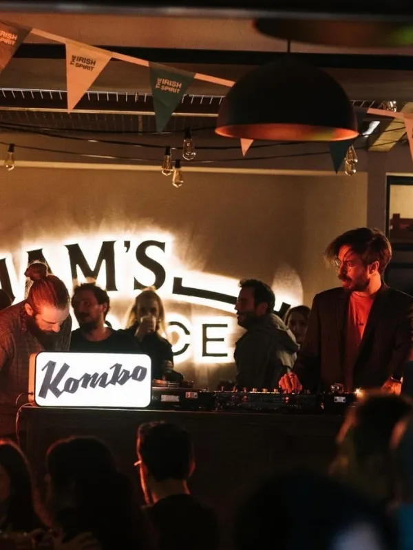 Loş ışıklı bir mekanda, üzerinde parlak Kombo tabelası bulunan bir DJ konsolunun başında müzik çalan bir DJ ve etrafında sosyalleşen kalabalık görünüyor; arka planda neon ışıklarla aydınlatılmış bir yazı, tavanda asılı duran Irish Spirit yazılı bayraklar ve sarkıt ışıklandırmalar dikkat çekiyor.