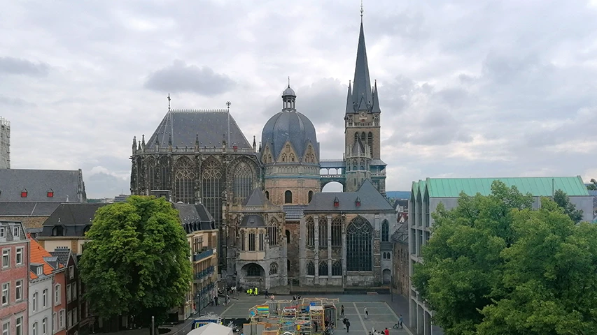 Aachen Katedrali'nin geniş ve çok katmanlı yapısı, gotik pencereler ve sivri bir çan kulesi ile büyük bir kubbeyi birleştirirken, gri bulutlu bir havada tarihi şehir meydanının ortasında görkemli bir şekilde yükseliyor ve önündeki canlı meydanda rengarenk stantlar ile hareketli insanlar dikkat çekiyor.