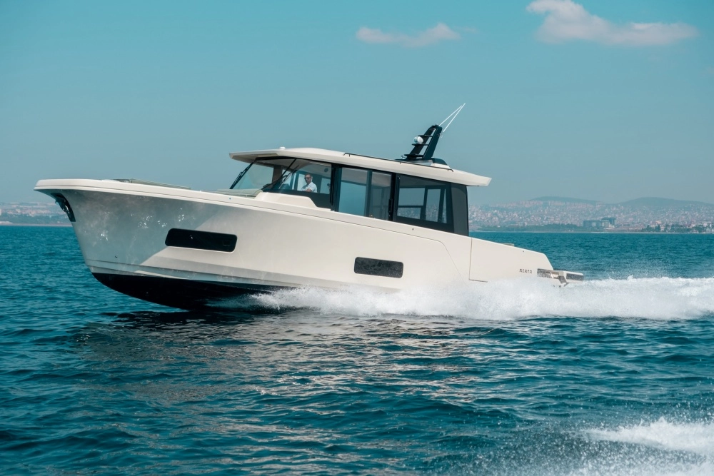 Anadolu Grubu'nun Türk yapımı modern motor yatı AIATA Wayfinder 50, masmavi sularda hızla ilerlerken denizde beyaz köpüklü bir iz bırakıyor ve arka planda siluet halinde bir şehir manzarasıyla dikkat çekiyor.