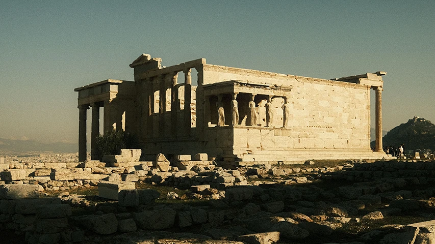Akropolis'te yer alan Erechtheion Tapınağı'nın, ünlü Karyatidler sundurması ve kısmen yıkık sütunlarıyla güneş ışıkları altında antik yapısını sergileyen bu görsel, Atina şehrinin üzerinde yükselen tarihi dokuyu vurguluyor.